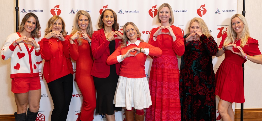 Group of women dressed in red making heart shapes with their hands standing in front of a banner with American Heart Association and ascension logos. Group of women dressed in red making heart shapes with their hands standing in front of a banner with American Heart Association and ascension logos.